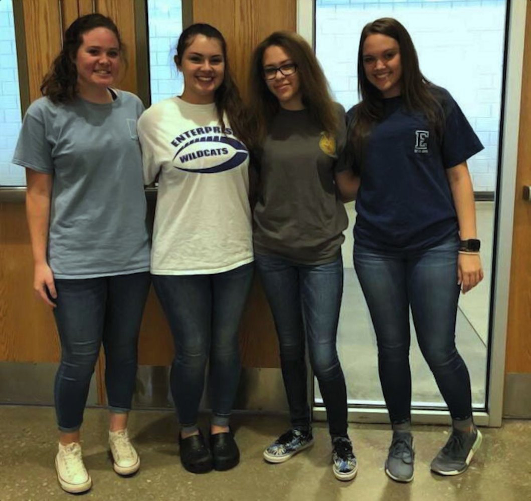 Vier studentes van de Enterprise High School staan naast elkaar binnenshuis, lachend naar de camera. Ze zijn nonchalant gekleed in jeans en T-shirts.