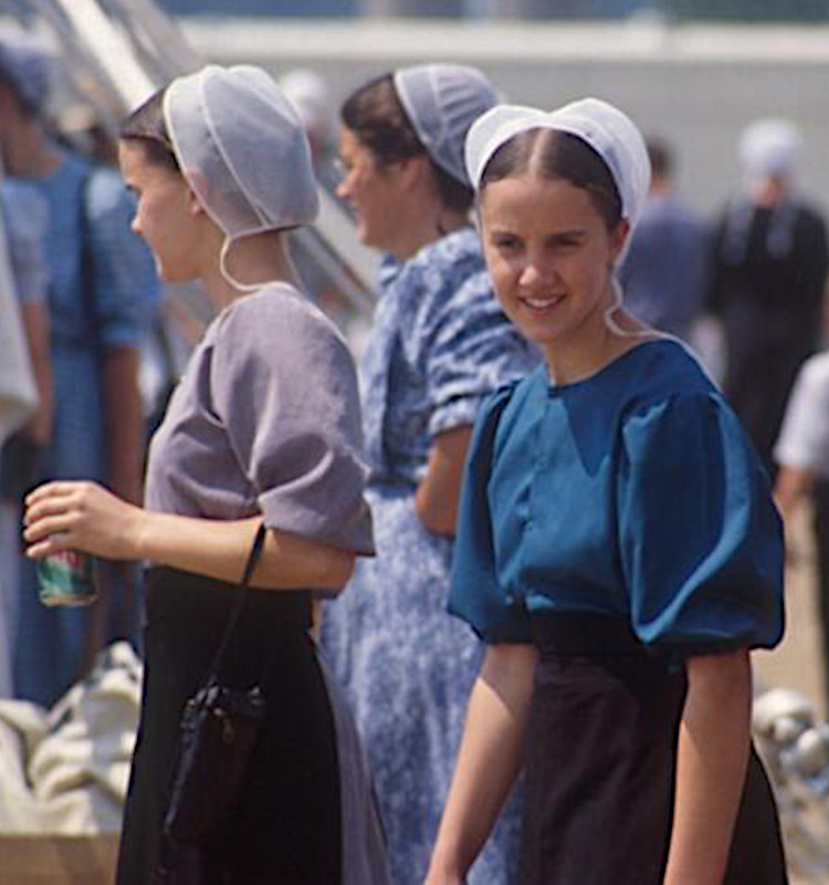 Twee jonge vrouwen, gekleed in traditionele Amish-kleding en witte mutsen, staan buiten te midden van een groep mensen in dezelfde kleding. Een van hen houdt een blikje drank vast.