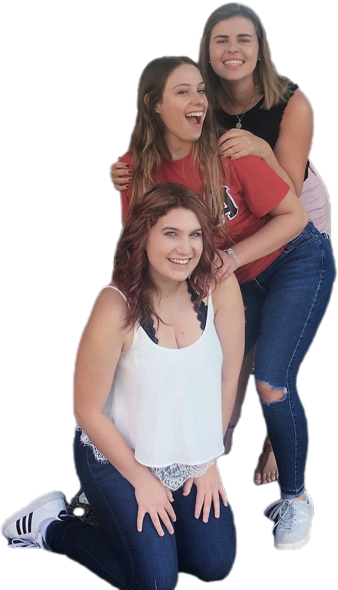 drie-vrolijke-meiden Drie jonge vrouwen poseren samen, glimlachend. Eén zit vooraan geknield, gekleed in een wit topje en een spijkerbroek. De twee achter haar staan, de een in een witte tanktop en spijkerbroek, de ander in een rood shirt en spijkerbroek.