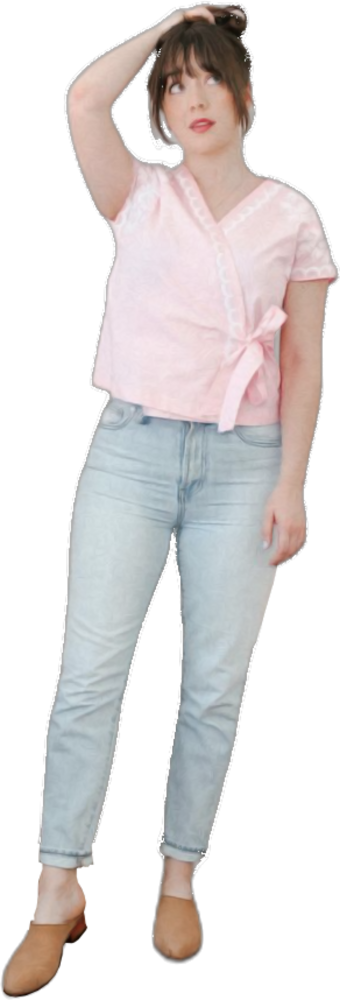Een dame staat met één hand naar het hoofd, gekleed in een roze blouse met korte mouwen, een strik en een lichtblauwe spijkerbroek, samen met bruine schoenen.