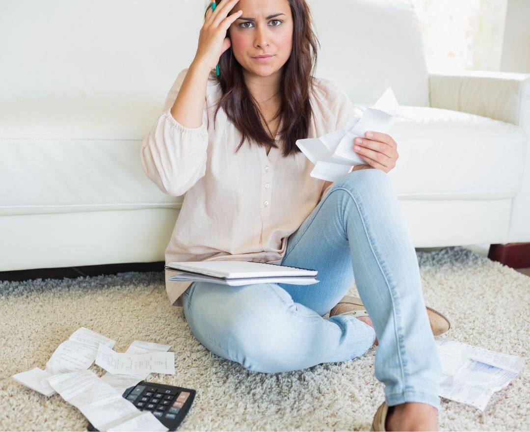 omgaan-met-geld Een vrouw die op een vloer met tapijt zit, ziet er gestresst uit terwijl ze verschillende papieren en bonnetjes in haar handen heeft, met een rekenmachine en nog meer verspreide papieren voor haar.