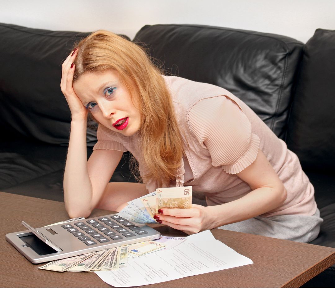 schulden-afbetalen Een vrouw met rood haar zit op een zwarte bank en houdt eurobankbiljetten vast. Ze ziet er gestrest uit, terwijl ze haar hoofd op één hand laat rusten, omringd door documenten, en een rekenmachine gebruikt.