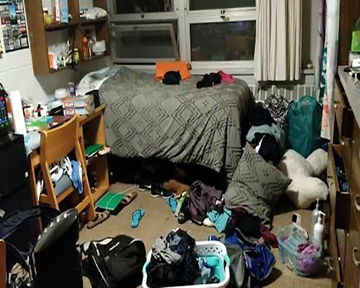 studentenkamer-rommel Een rommelige kamer met kleding, tassen en diverse spullen verspreid over de vloer. Een bed met een grijze deken is gedeeltelijk zichtbaar, samen met een bureau, een stoel en planken vol spullen.