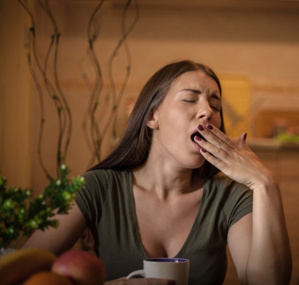 telaat-naar-bed Een vrouw zit aan een tafel met een mok, geeuwend met gesloten ogen en hand voor haar mond. Ze draagt een groen shirt en voor haar staan planten en fruit op tafel.