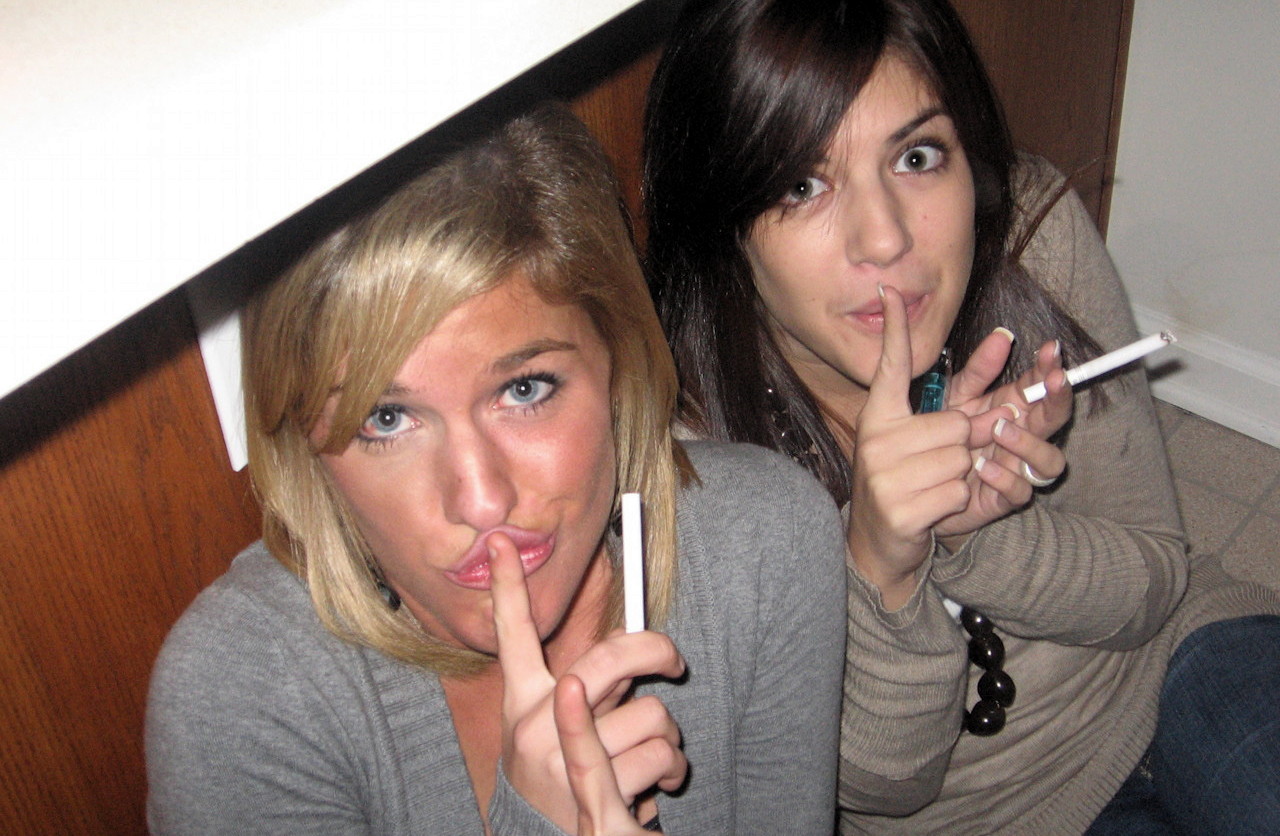 Twee vrouwen zitten op de grond onder een toonbank, met sigaretten in hun handen, met de vingers tegen hun lippen in een zwijgend gebaar.
