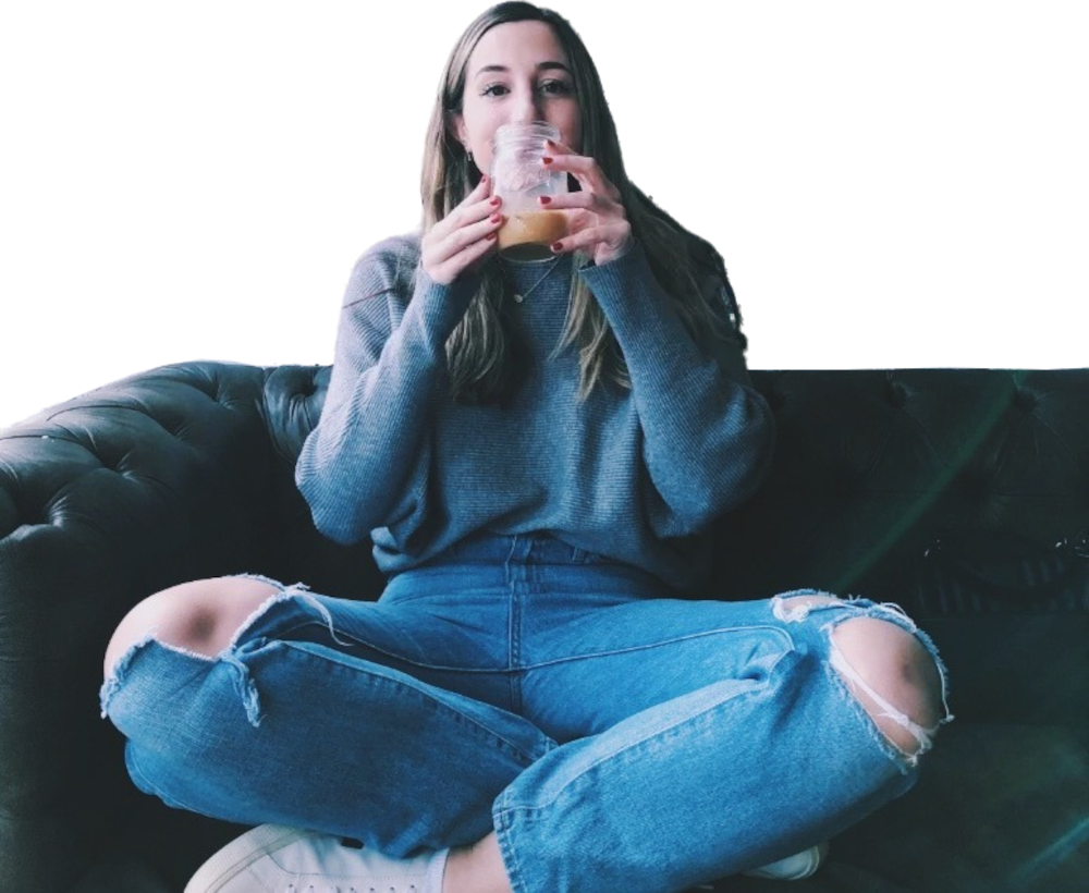 Een studente in een grijze trui en een gescheurde spijkerbroek zit met gekruiste benen op een bank en drinkt uit een glazen pot.