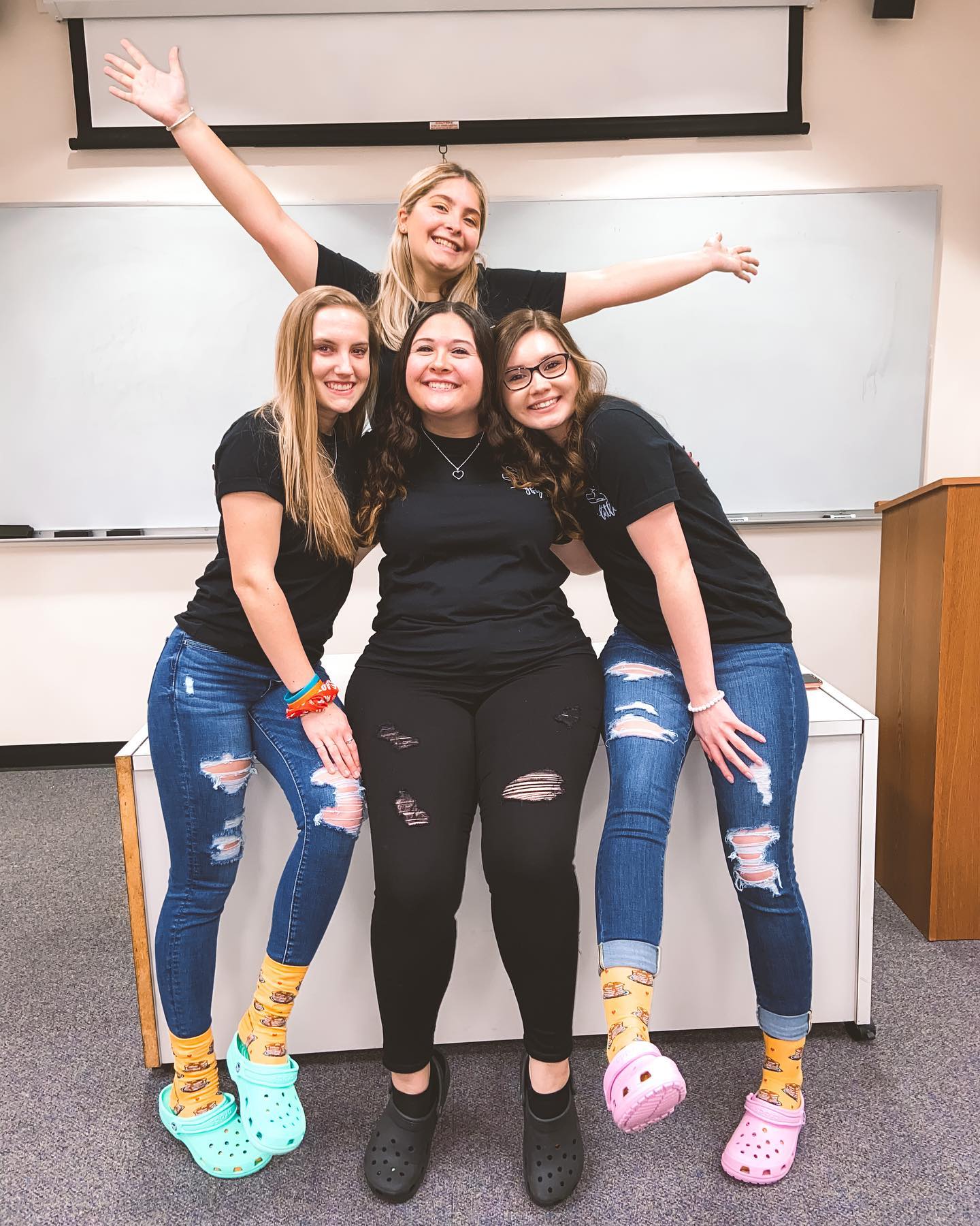 Vier studentes poseren lachend voor een whiteboard. Drie staan achter een zittende persoon, met kleurrijke sokken en Crocs, gescheurde jeans en zwarte shirts.