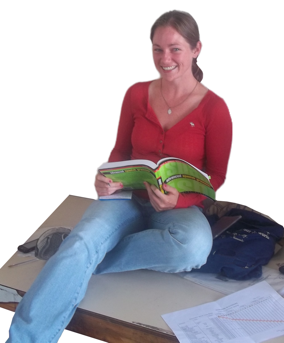 Een studente in een rood topje en jeans zit met gekruiste benen op een tafel, glimlachend terwijl ze een open boek vasthoudt. Papieren en een rugzak liggen in de buurt.