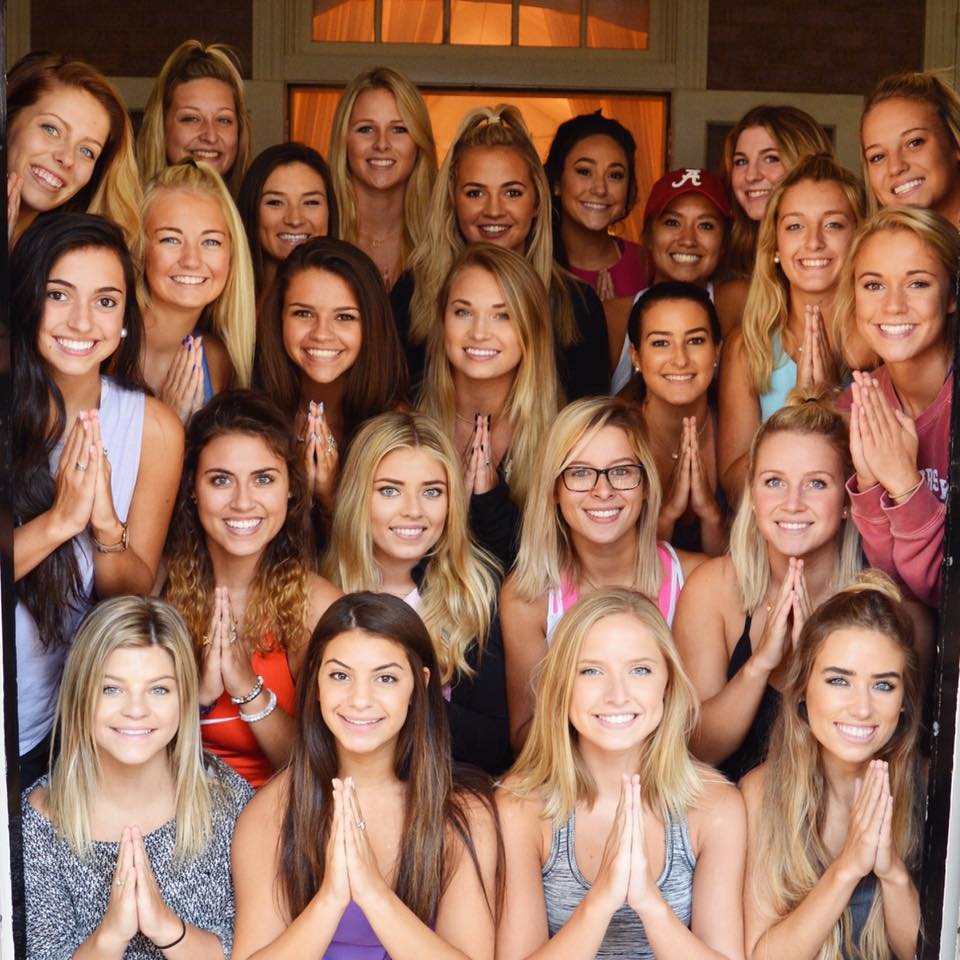 Een groep van twintig leden van een sorority poseert samen voor de deur van hun huis. Ze glimlachen en houden hun handen in gebedshouding, staand en zittend dicht op elkaar in rijen.