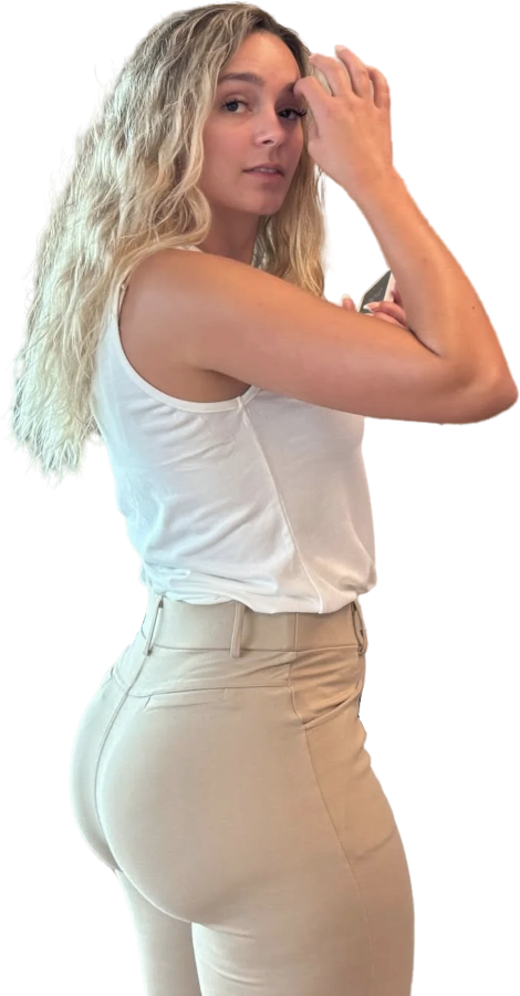 Een vrouw, gezien van haar rechterkant. in haar linkerhand heeft ze een smartphone, met haar rechterhand veegt ze de haren uit haar gezicht. Ze draagt een lichtbeige broek die haar billen goed laten uitkomen. Ze kijkt een beetje provocerend in de camera.
