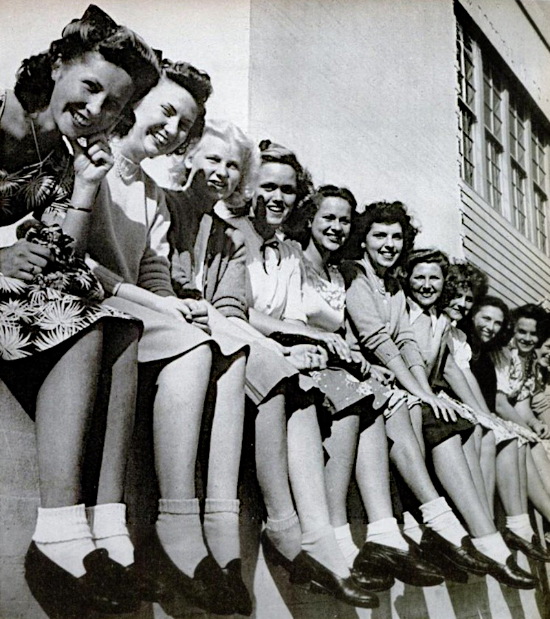 Een foto uit de jaren vijftig met een groep studentes die naast elkaar zitten op een muur bij een schoolgebouw.