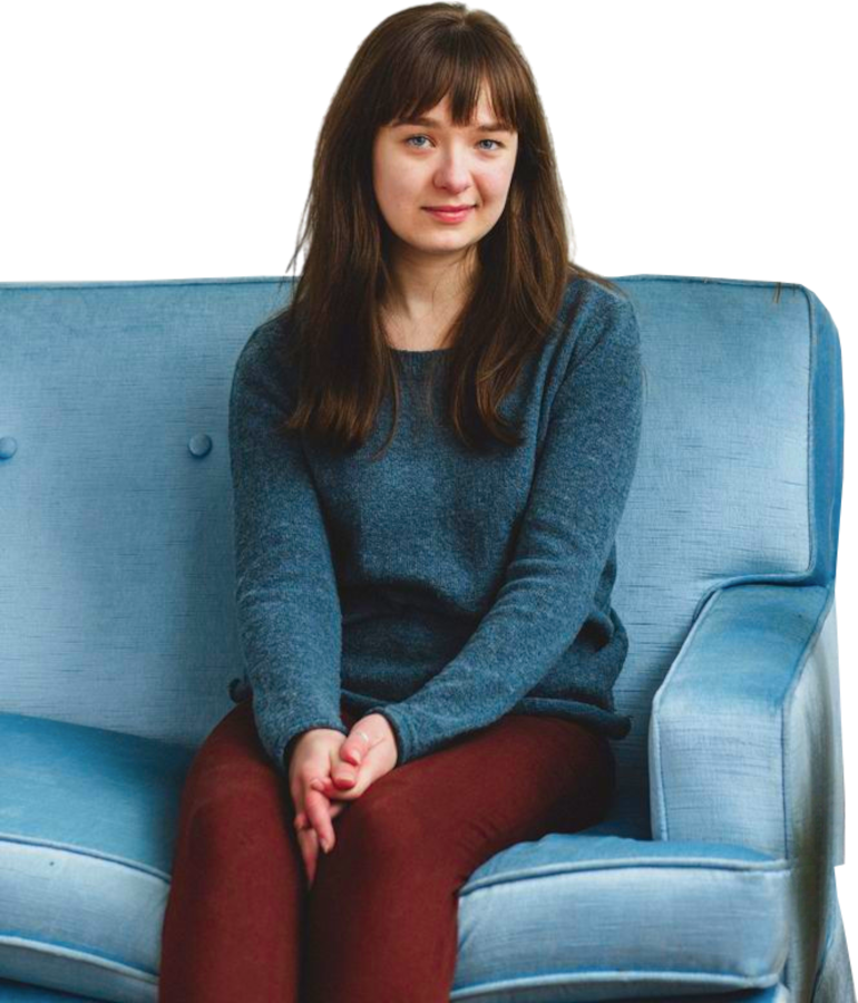 Een vrouw zit op een lichtblauwe sofa. Ze is gekleed in een een blauwe truie en een bordeaux rode broek. Ze kijkt vol verwachting in de camera.