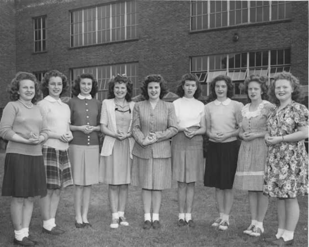 Een vintage foto van een groep van 9 jonge vrouwen voor een industrieel gebouw.